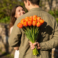 A man hides a bunch of orange tulips tied with garden twine behind his back to surprise a smiling woman, outdoors in the sunshine. Laura Florals