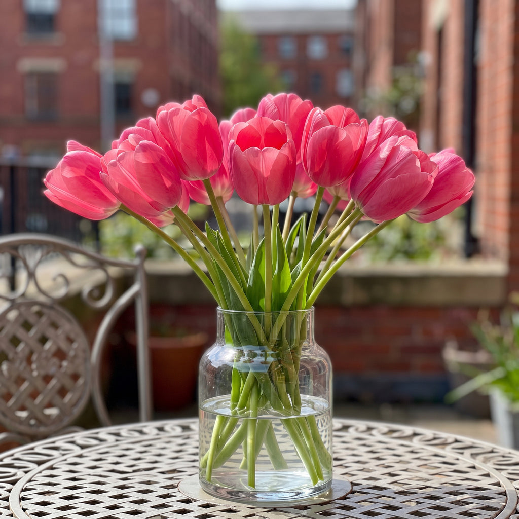 A glass vase of vibrant Tulip 'Pink Impression' blooms on a bistro table on a sunny Manchester, UK, balcony
