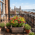 An upcycled wooden planter box filled with a colourful mix of zinnia flowers from Laura Florals flower planting kits sits on a wooden balcony deck, surrounded by potted herbs, a copper watering can, and a black wrought-iron railing. In the background, a scenic view of Liverpool and harbour is visible under a blue sky.